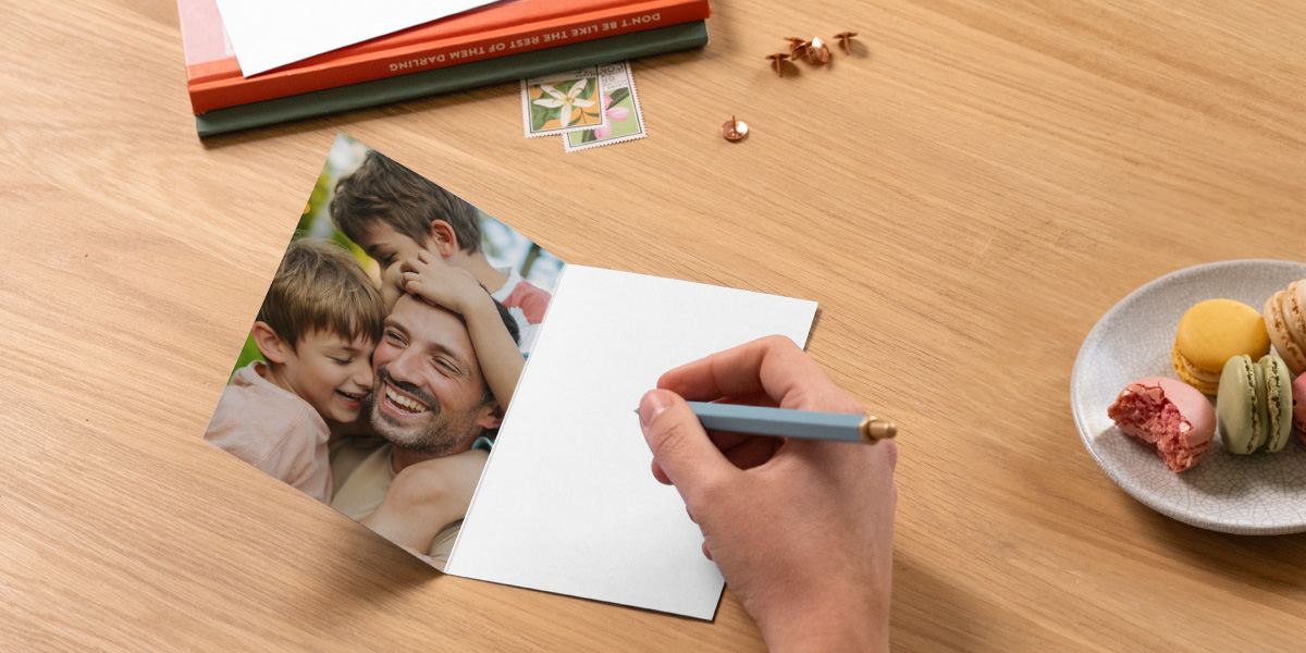 An open personalised Photobox Father’s Day photo card shows a father with his two sons, with a pen poised to write a message beside books and a plate of macarons, inspiring what to write in a Father’s Day card.