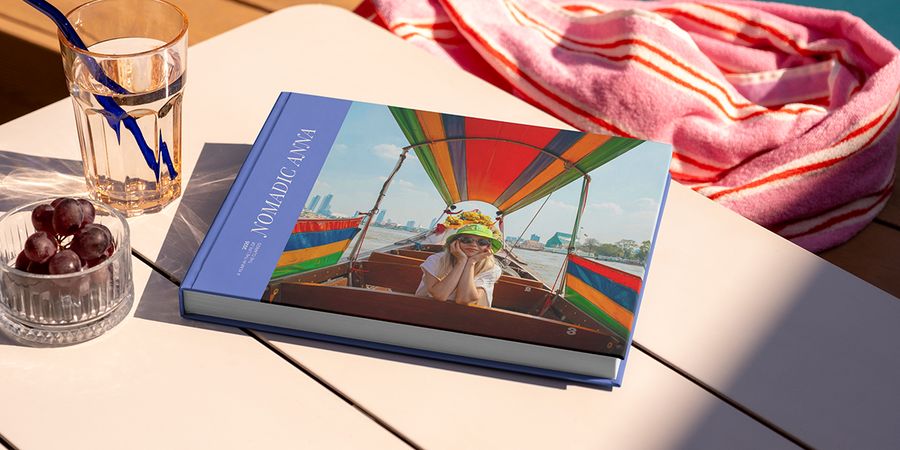 A large landscape travel photo book titled 'Nomadic Anna' sits on a table, featuring a photo of a girl on her travels on the cover, illustrating Photobox's travel photo album title ideas.