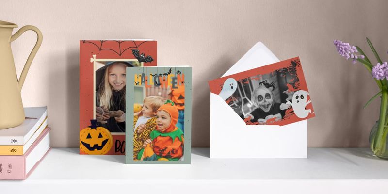 Three personalised Halloween cards sit side by side on a mantlepiece, featuring a little girl dressed as a witch, a little boy dressed as a pumpkin, and a little girl dressed as a ghost surrounded by ghost illustrations, introducing Photobox's guide to creating killer Halloween party invites.