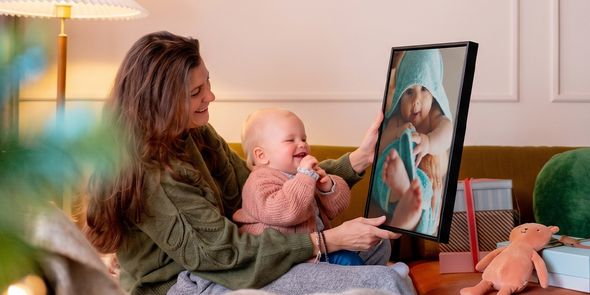 Woman with a baby on her lap in a living room, holding up personalised wall art of the baby while the baby looks at it and giggles, illustrating Photobox’s baby’s first Christmas photo gift ideas.