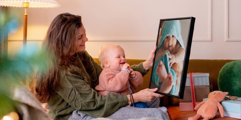 Woman with a baby on her lap in a living room, holding up personalised wall art of the baby while the baby looks at it and giggles, illustrating Photobox’s baby’s first Christmas photo gift ideas.