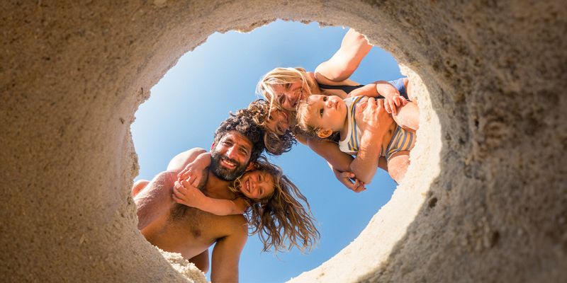 A family of four on the beach – mum, dad, daughter and baby boy – look into a hole dug in the sand, capturing a playful summer moment and inspiring beach photos and beach photography ideas.