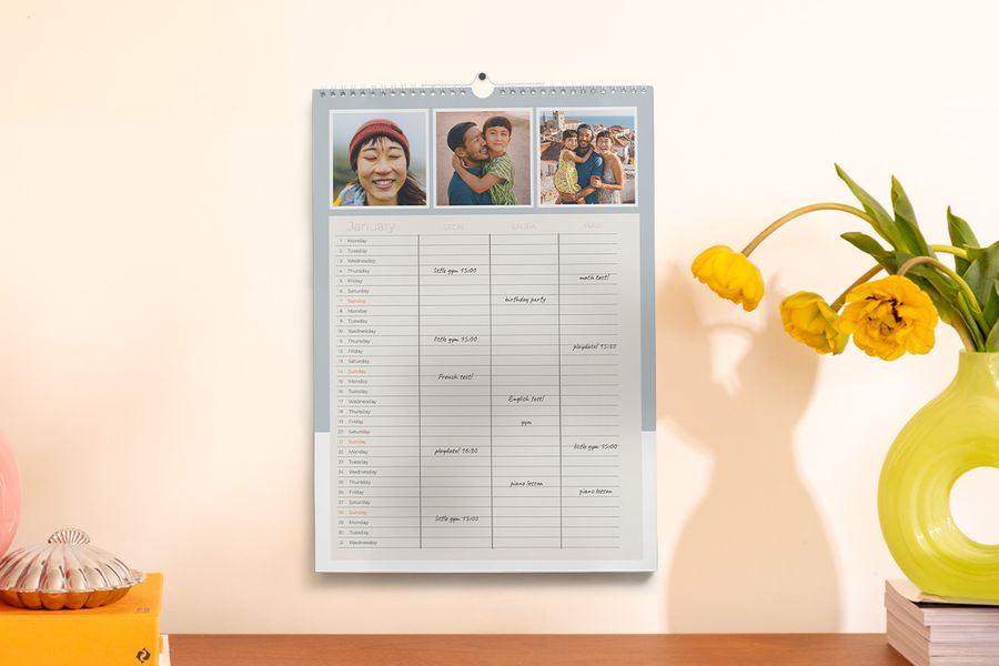 A family planner hangs on a wall next to a vase and some books, illustrating Photobox's tip to stay organised for the school year with a personalised planner.