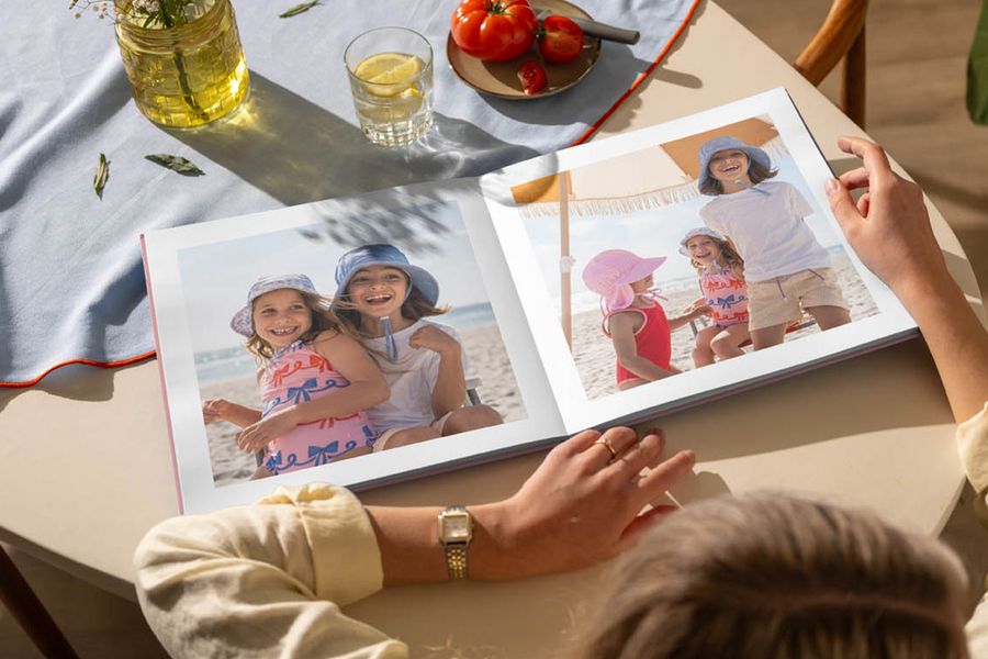 An open photo book on a kitchen table features images of young girls having fun at the beach, illustrating Photobox's tip to look back on summer memories with a personalised photo book.