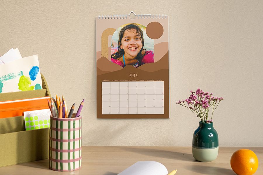 A personalised calendar showing September and featuring a photo of a young girl hangs above a desk with a pencil pot, a vase of flowers and an organiser holding art and stationery supplies, illustrating Photobox's tip to create a focused study space for the school year.