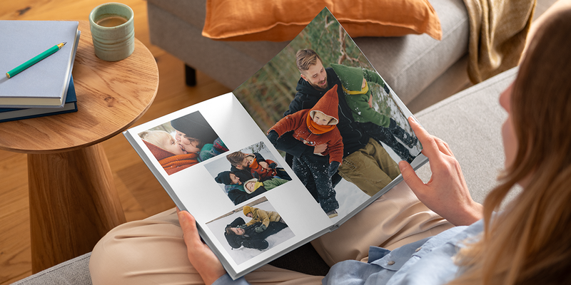 A woman looking at an open premium layflat photo book from Photobox featuring family photos in the snow, showing a high-quality personalised photo album.