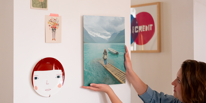 A woman places personalised wall art on her wall next to an ornamental plate and some photo prints, introducing Photobox's step-by-step guide to hanging wall art with hanging kits.