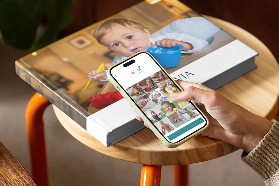 Hand scrolling through baby pictures on a smartphone with a first photo album from Photobox on a wooden table in the background.