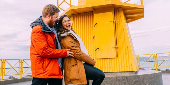 A happy couple laughing and having fun in front of a yellow lighthouse while searching for what to write for Valentine's Day and valentines day ideas for their Photobox gifts.