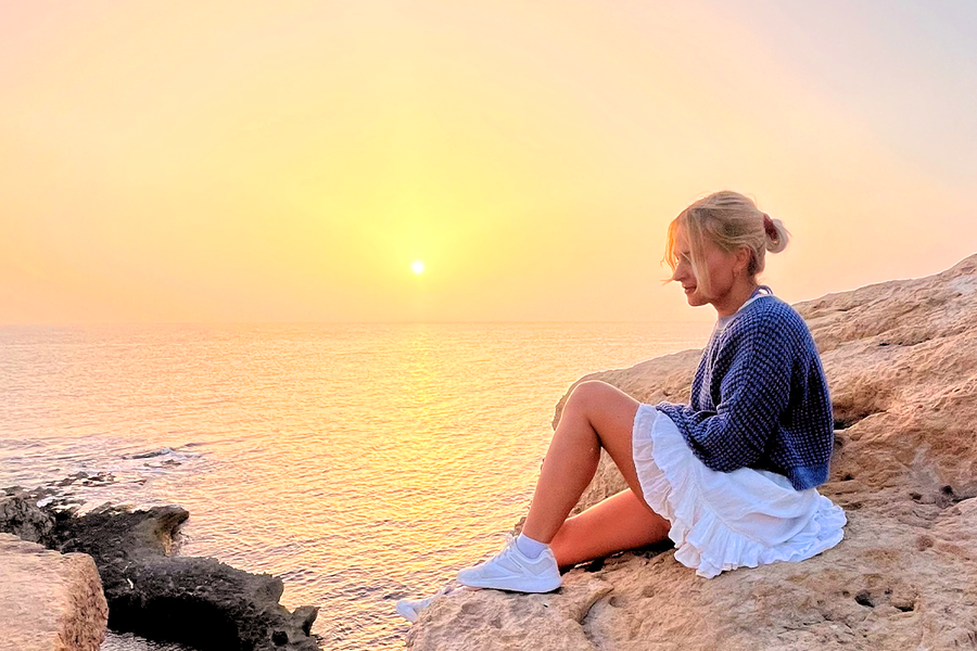 A woman sitting on rocks by the sea with the sun setting behind her, illustrating how golden hour light can create warm and beautiful photos.