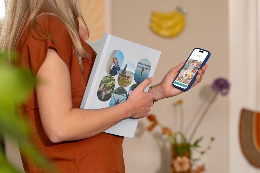 A woman holds a closed Photobox holiday photo album with family travel photos on the cover while looking at travel images on her phone, illustrating how to choose the right photos for a personalised travel photo book.