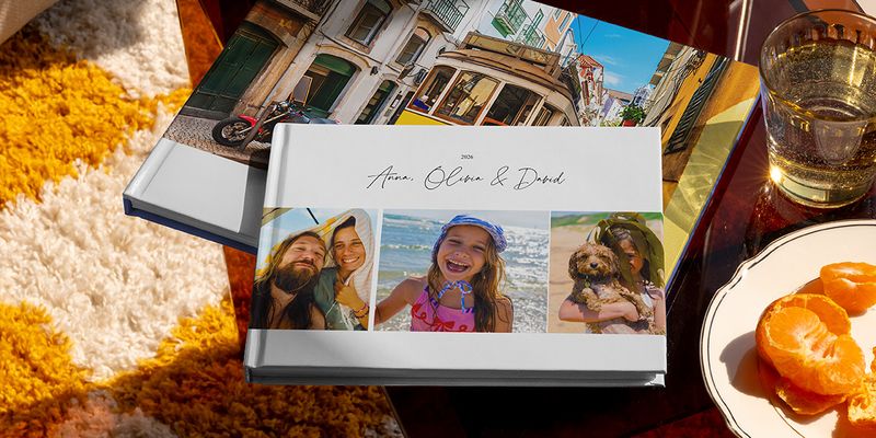 Two different Photobox travel photo books are displayed together, including a landscape holiday photo book with family travel photos on the cover and another with a tram holiday shot, showing different personalised travel photo book ideas.
