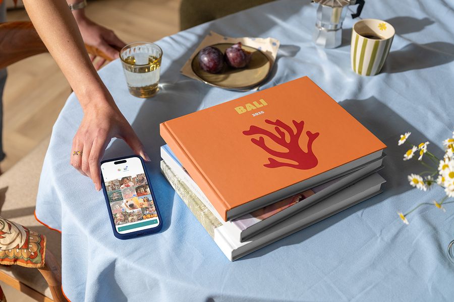 A stack of different Photobox travel photo books sits beside a hand holding a phone with a holiday photo ready to upload, with the top holiday photo book labelled Bali 2026 and showing coral on the cover, illustrating how to create a personalised travel photo book.
