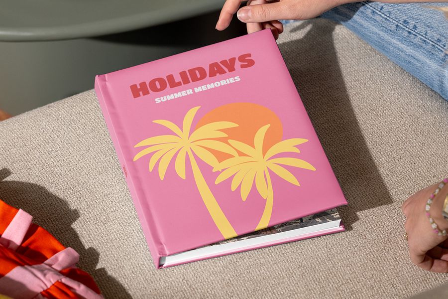 A closed Photobox travel photo book called “Holidays, summer memories” with illustrated palm trees on a pink cover is held by hands ready to open it, inspiring how to structure a holiday photo album and tell your travel story.