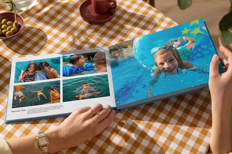 An open Photobox holiday photo book shows family summer photos playing in the water, with one full-page spread and another page titled Marseille 2026 with a caption and description, illustrating how captions bring a personalised travel photo book to life.