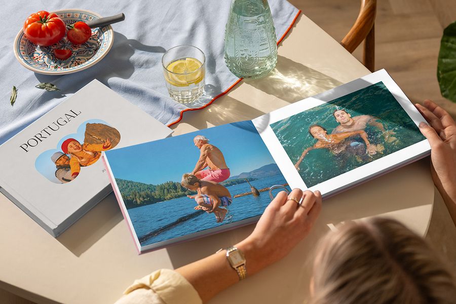 Two different Photobox travel photo books are shown, with a woman looking through an open holiday photo book filled with family photos by the sea, alongside a closed Portugal travel photo book, illustrating a personalised holiday photo album full of travel memories.