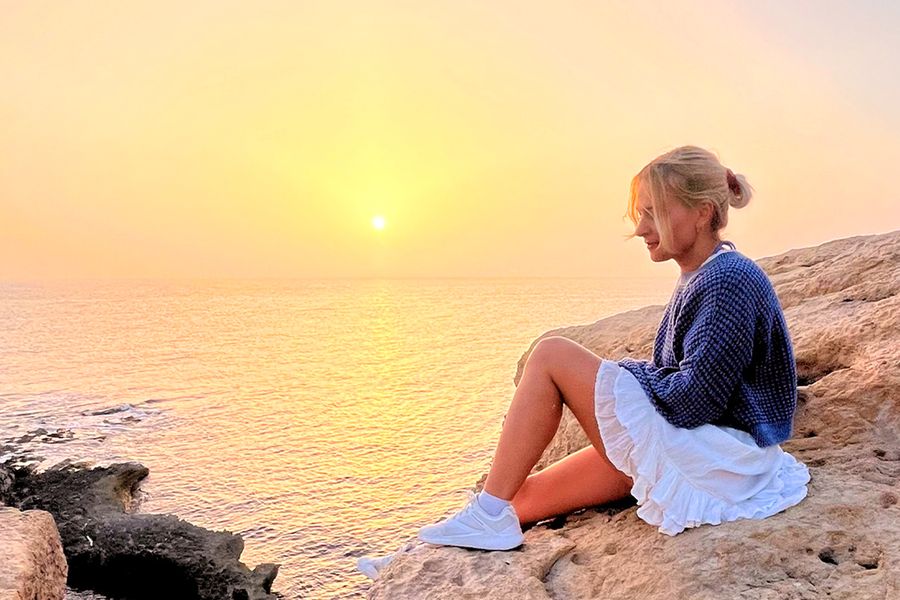 A woman sits on rocks looking out at the sea and sunset, capturing the spirit of Photobox's adventure staycation ideas.