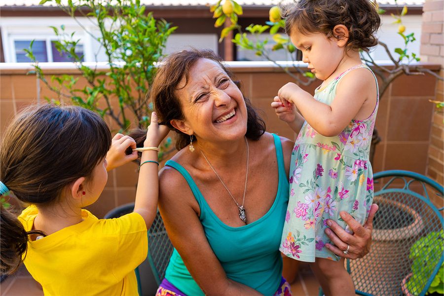 A woman sits on a balcony while two young girls play with and style her hair, a lighthearted moment from Photobox's family staycation ideas.