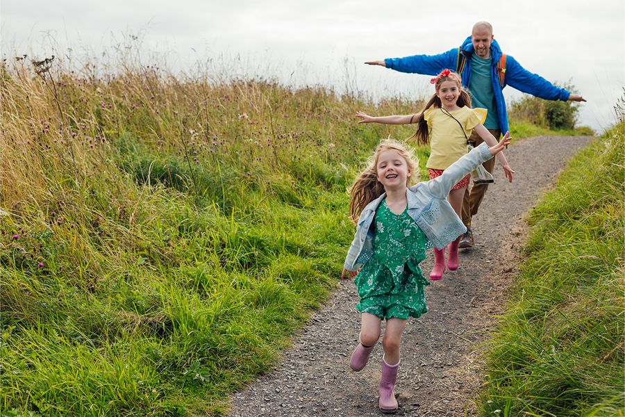 A father and his two daughters play and run through a field, reflecting Photobox's staycation ideas near nature.