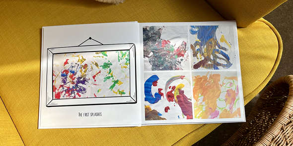 An open children's photo book rests on a yellow armchair, showing young children's early splash paintings on the left page and four different paintings on the right, illustrating Photobox's ideas for a children's photo album.