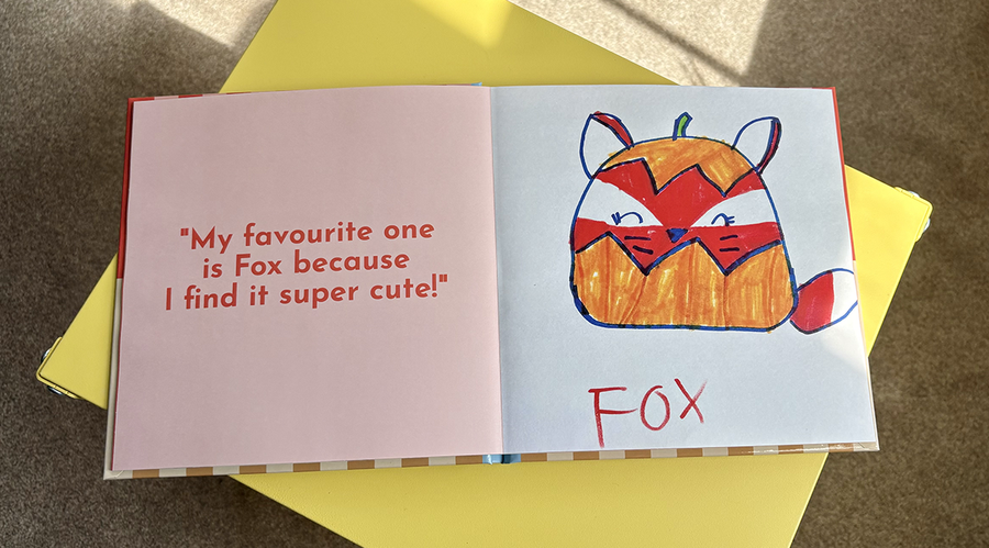 An open children's photo book shows a caption and quote from a child explaining her artwork on the left, alongside her corresponding drawing of a cartoon fox on the right, illustrating Photobox's tips for adding text to a children's photo album.