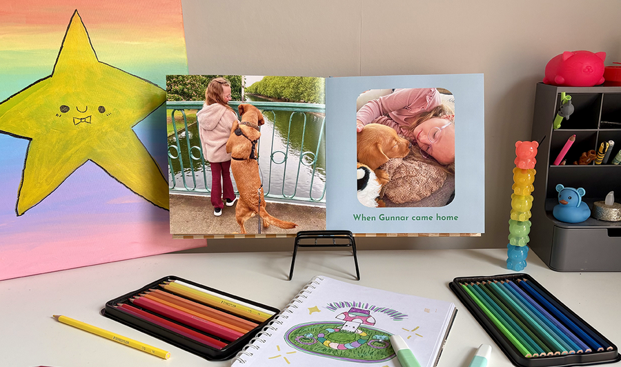 An open children's photo book rests on a child's desk surrounded by colouring pencils and artwork, showing a photo of a girl and her dog on the left page and another captioned image on the right, illustrating Photobox's tips for mixing photos and artwork in a family photo book.