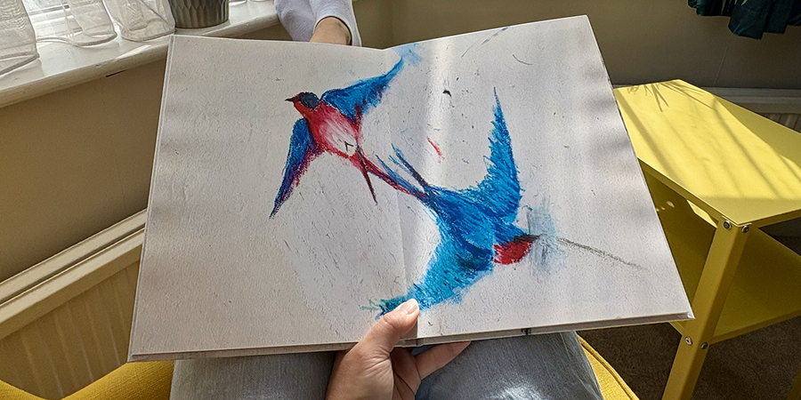 An open children's photo book with a full-page spread of a bird drawing rests on a woman's lap as she sits in an armchair, illustrating Photobox's steps for creating a family photo book from a child's drawings.