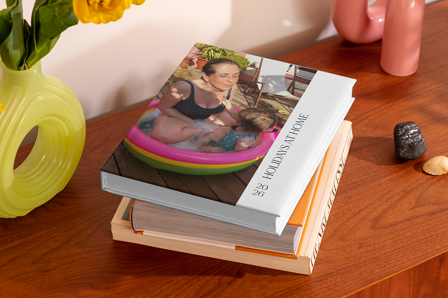 A stack of photo books features a top book showing a woman and a young child in a paddling pool, titled 'Holidays at Home', illustrating Photobox's staycation photo album title ideas.