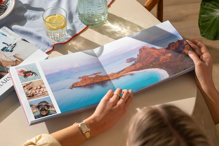 An open travel photo book on a table shows a double-page spread of a beautiful beach landscape alongside three smaller summer photos, illustrating Photobox's Travel Chapter design from the new Summer Travel Series.