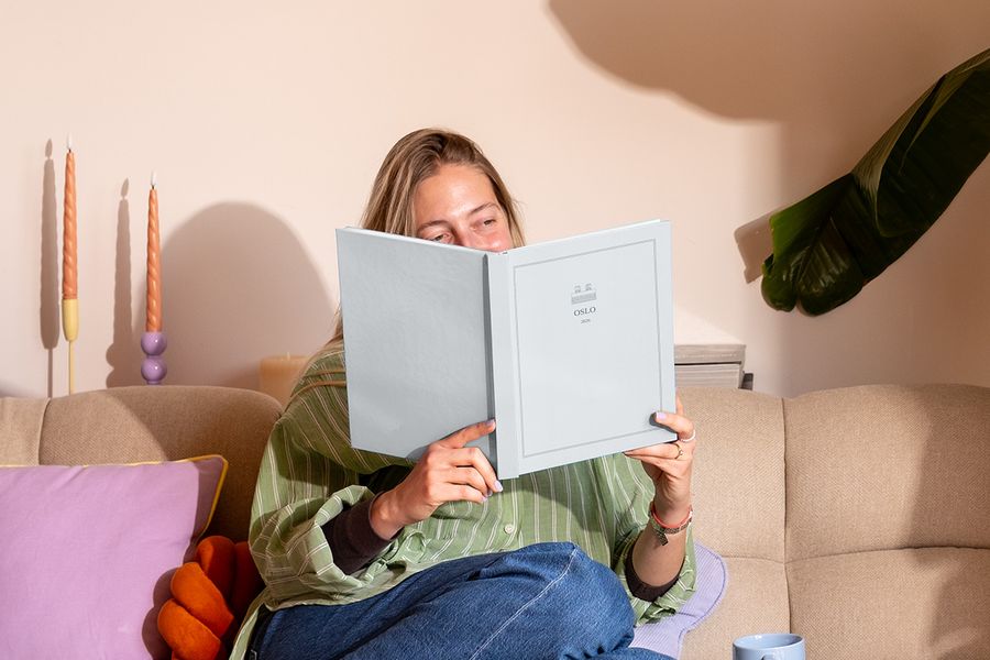 A woman sits on the sofa next to a colourful cushion, looking at a personalised travel photo book titled 'Oslo', showcasing Photobox's new Summer Travel Series designs.