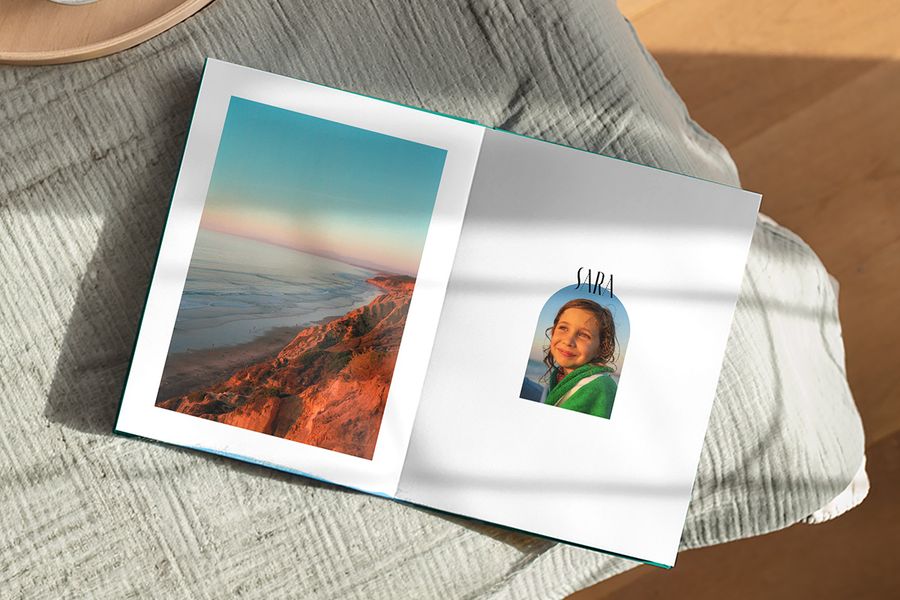 An open photo book shows a beautiful landscape on one page and a picture of a girl captioned 'Sara' on the other, illustrating Photobox's Artisan Masking design with playful frames inspired by vintage postcards and postage stamps.
