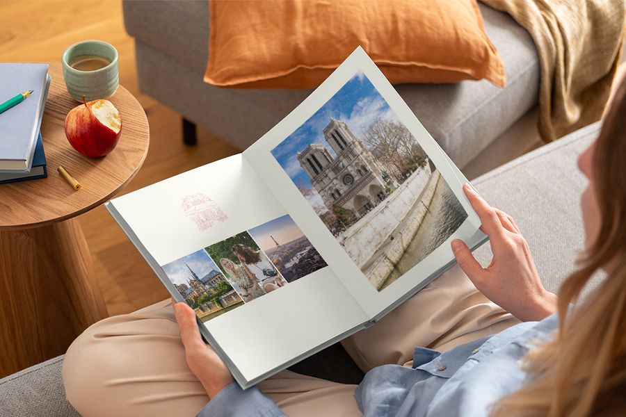 A woman holds an open photo book on her lap showing different travel shots, illustrating Photobox's Graphic Getaways design inspired by vintage travel posters and illustrated city maps.