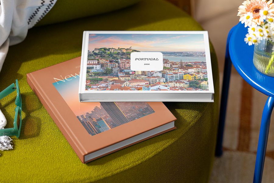Two personalised photo books are stacked on a stool, the top titled 'Portugal' with a picture of Portugal, and the one beneath titled 'New York' with a picture of the city, showcasing Photobox's customisable Summer Travel Series designs.