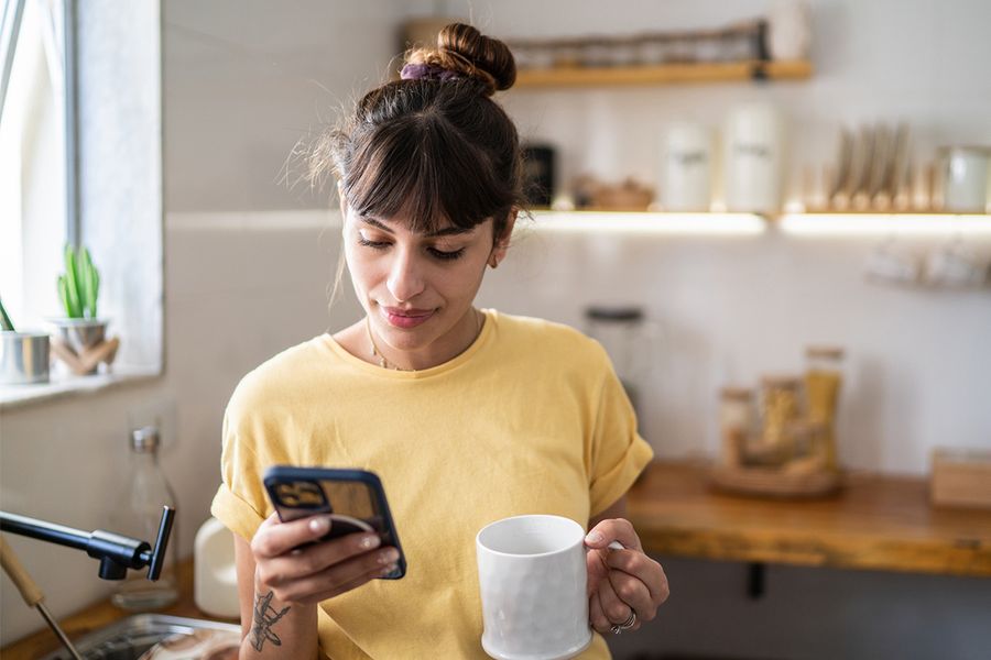 A woman in her kitchen holds a mug while looking through her phone, illustrating Photobox's tips for building photo organising habits that actually stick.