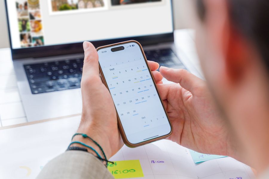 Someone looks at the calendar on their phone while working on a photo book on their laptop in the background, illustrating Photobox's simple monthly photo maintenance routine.