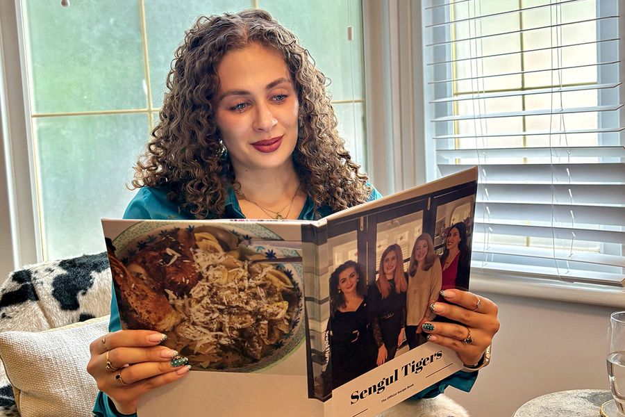 Sarah, who works at Photobox, sits in a cow-print chair looking at her personalised family recipe book, with the cover showing four women, including Sarah, illustrating how a recipe photo book brings together family recipes and memories.