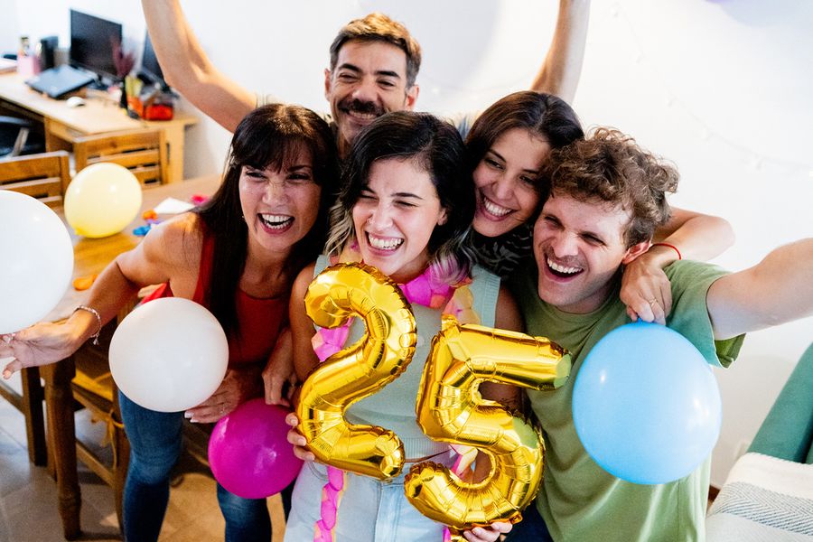 A group of young people crowd around '25' age balloons, smiling and posing for a photo, illustrating Photobox's tips for capturing milestone birthday moments.