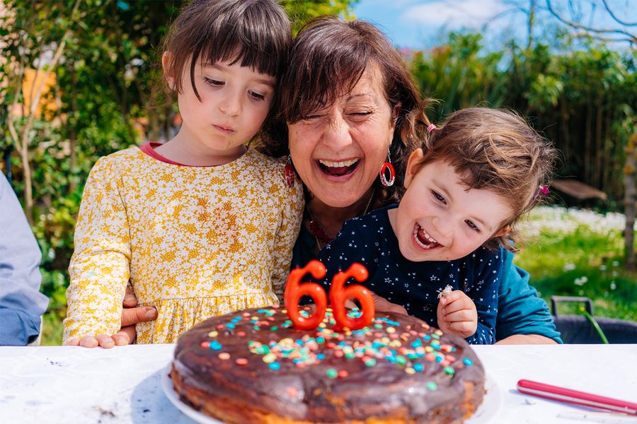 A woman smiles beside her birthday cake with candles showing '66', flanked by two smiling children, illustrating Photobox's tips for capturing multi-generational birthday moments.