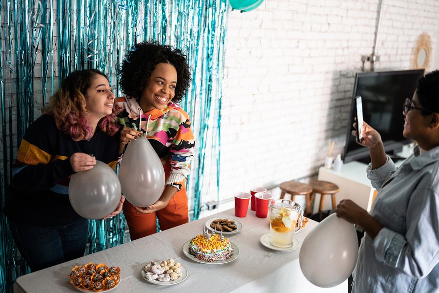 Two girls pose in front of a tinsel photo backdrop, holding balloons beside a snack table filled with party food and a birthday cake, illustrating Photobox's tips for photographing party decor and food layouts.