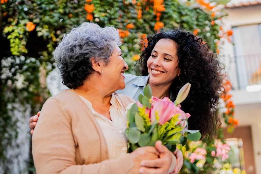 A young woman smiles at her mum, who is holding flowers, illustrating Photobox's ideas for what to write in an anniversary card for parents.