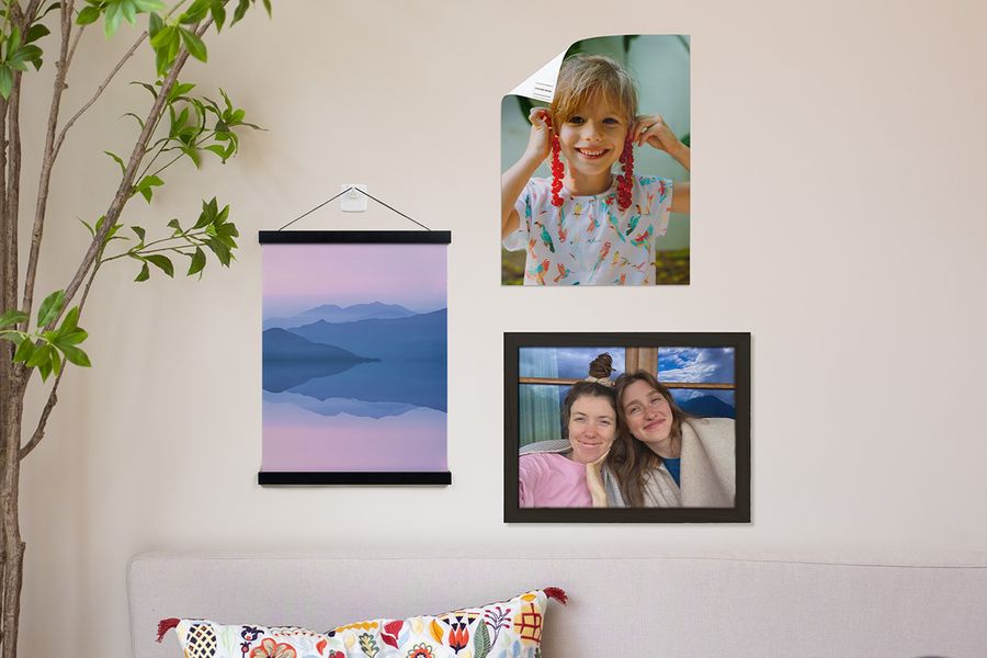 Different Photobox wall art hanging options are shown in a home, including a personalised poster of a young boy attached with adhesive strips, a personalised landscape poster hung with a wooden hanger and adhesive hook, and a framed picture of two smiling girls.