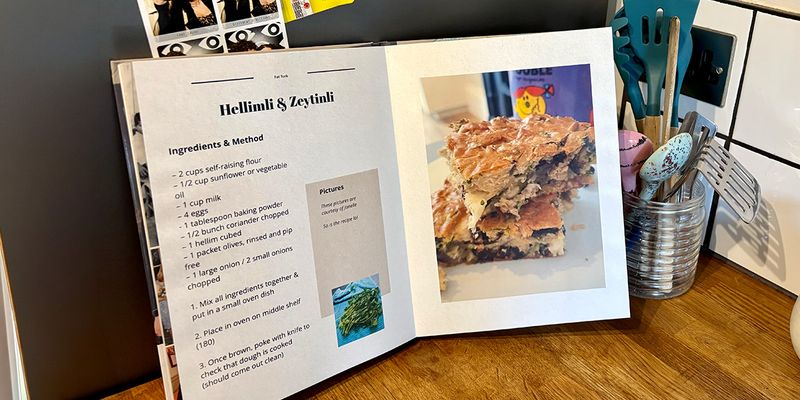 An open personalised recipe book sits in a kitchen setting, with a jar of spoons and cooking utensils in the background, introducing Photobox's guide to making your own cookbook.