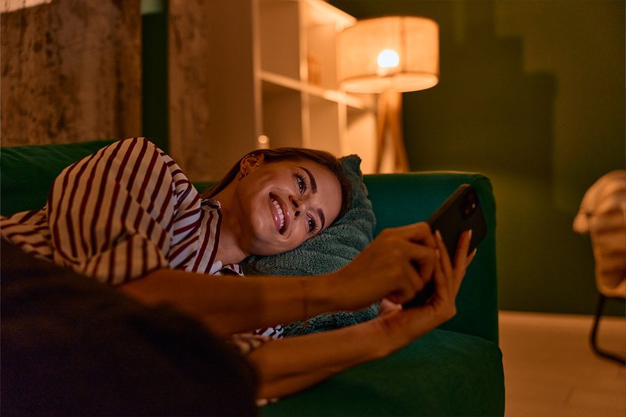 A woman lies on a dark green sofa looking at her phone in a living room, lit by a lamp for cosy lighting, illustrating Photobox's tips for hygge lighting at home.
