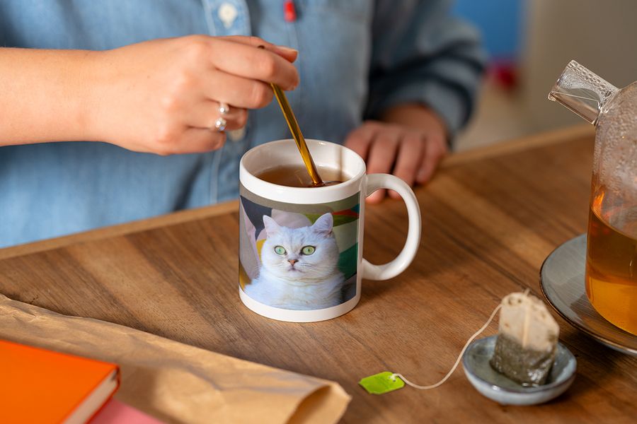 Someone stirs a spoon in a personalised photo mug featuring a white cat photo while making a cup of tea, illustrating Photobox's tip to finish a hygge reading nook with a personalised mug.