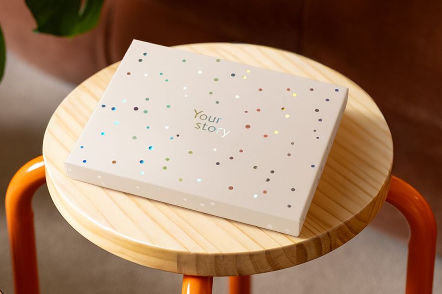 A Photobox presentation box rests on a wooden stool, decorated with silver holographic circles and the words 'Your Story', illustrating the matte ivory design with a touch of holographic shine.