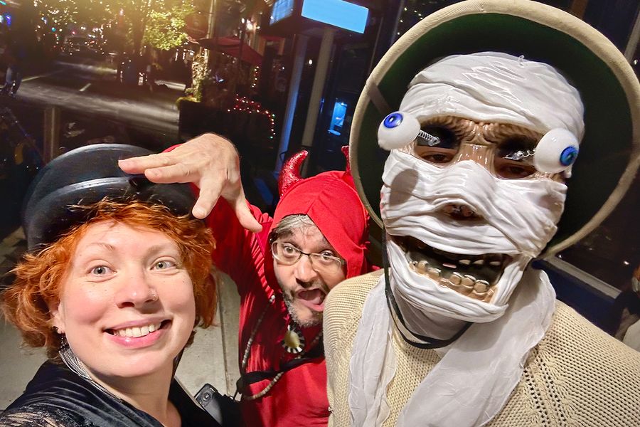 Three adults in Halloween fancy dress on the street, one in a skeleton mask with popping eyes, one dressed as a devil and one woman wearing a black hat, illustrating Photobox's Halloween caption ideas for costumes.