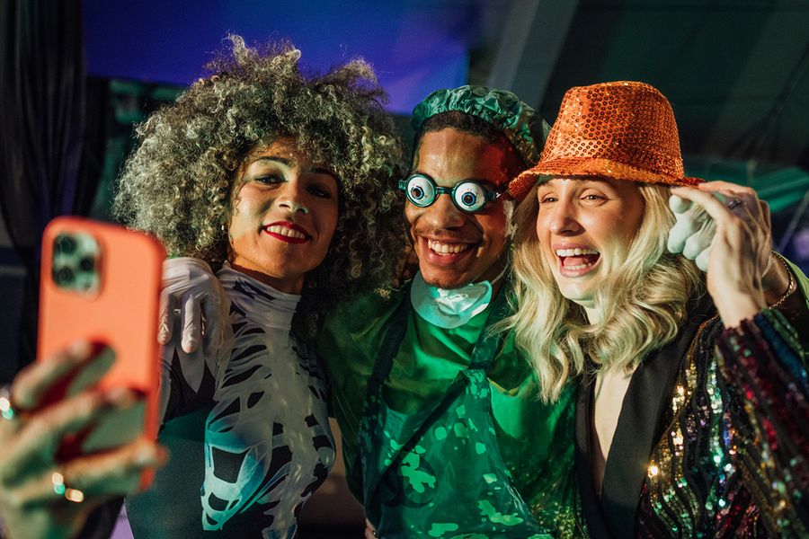 Three adults pose for a selfie at a Halloween party, one woman in a skeleton costume, a man dressed as a scary surgeon with funny glasses and a woman in a sequinned hat, illustrating Photobox's group Halloween captions.