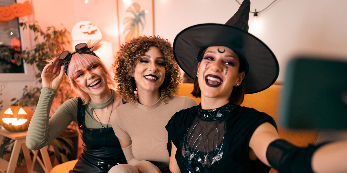 Three women in fancy dress at a Halloween party, a witch, an animal and a cyberpunk, pose for a selfie on their phone, introducing Photobox's 105 Halloween Instagram captions.