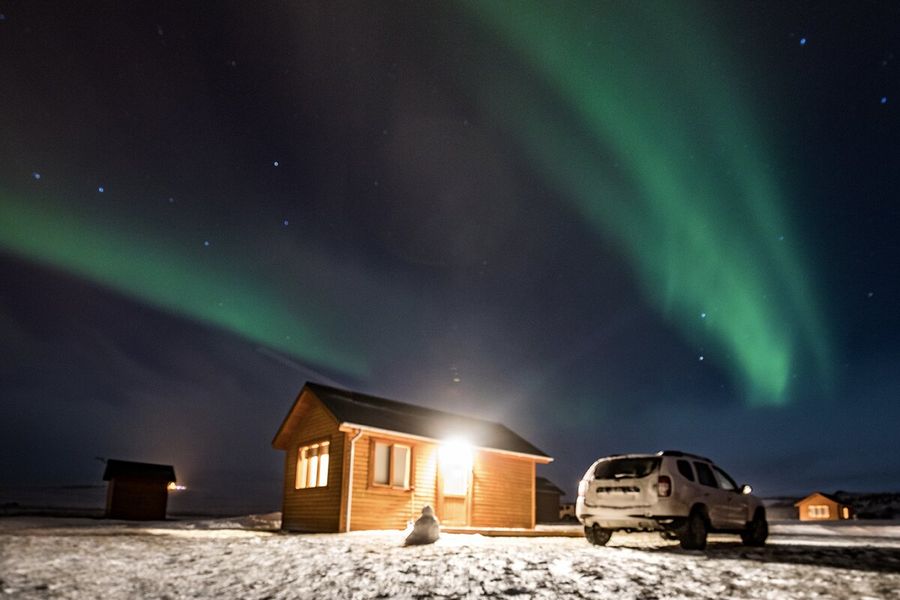 A cabin with a light shining out next to a car in a very snowy landscape, with green northern lights in the sky above, illustrating Photobox’s guide to what the northern lights are and what causes aurora borealis.