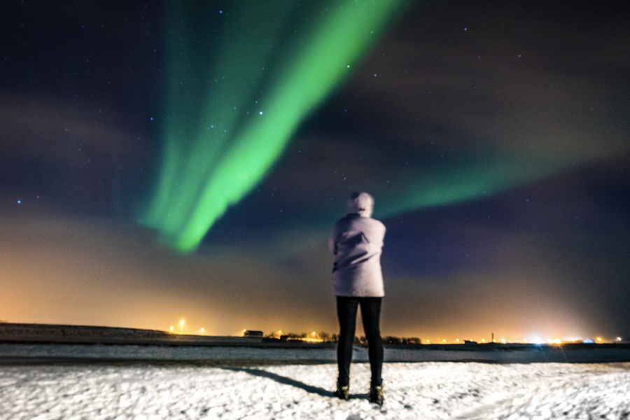 A person standing in a snowy scene looking up at green northern lights in the sky, illustrating Photobox’s tips on how to photograph aurora borealis with a smartphone.